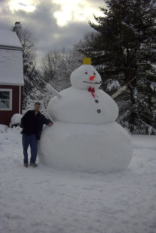 A man standing beside a snowman in the woods, with a giant snowman towering in the background