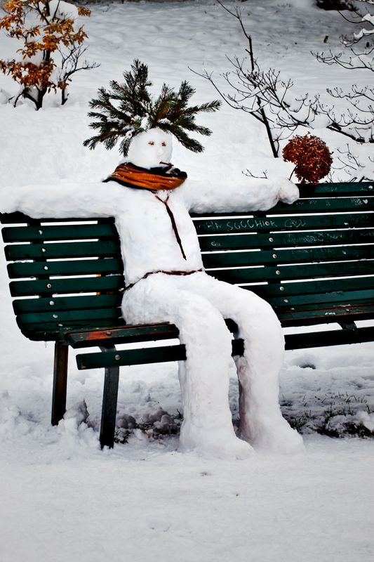 A snowman sitting on a bench in the snow, with a man relaxing beside it