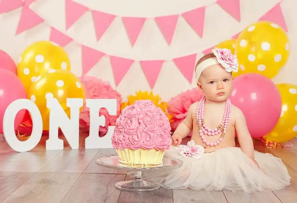 A one-year-old girl in a tutu and pink dress poses in front of a cake with a pink cupcake for her First Birthday Bash