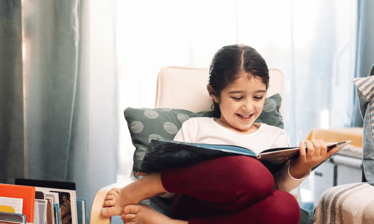 A young girl engrossed in a book while sitting on a chair. Image: How to Find Sports Chapter Books for 3rd Graders