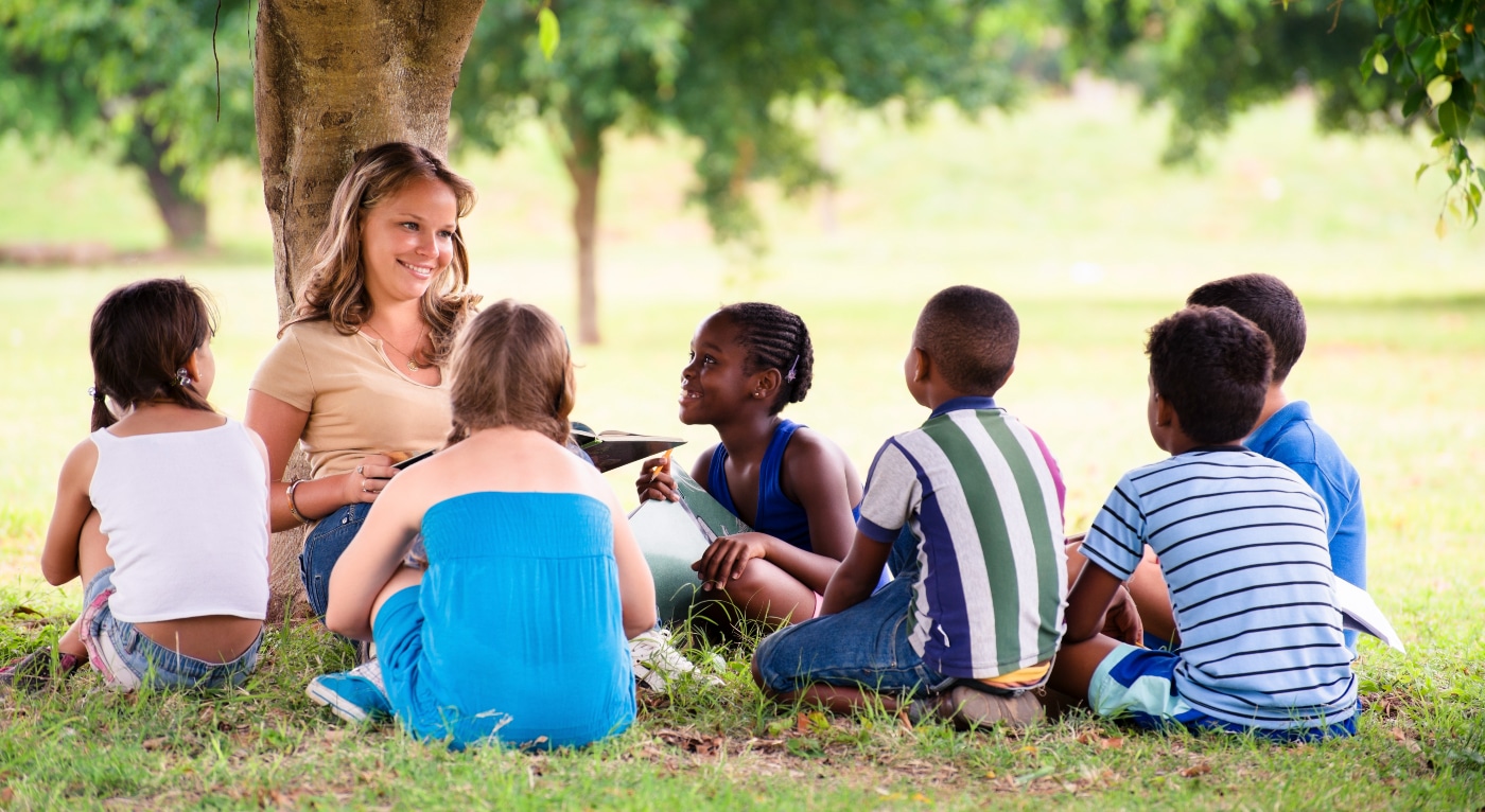 A woman sitting under a tree with children, embracing the importance of outdoor learning in a child's development