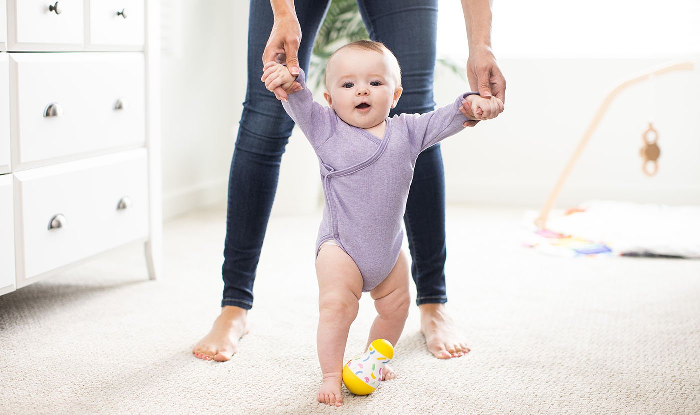 A woman supports a baby's hand while they stand on the floor. Milestone: Month 10-12: First Steps and Words