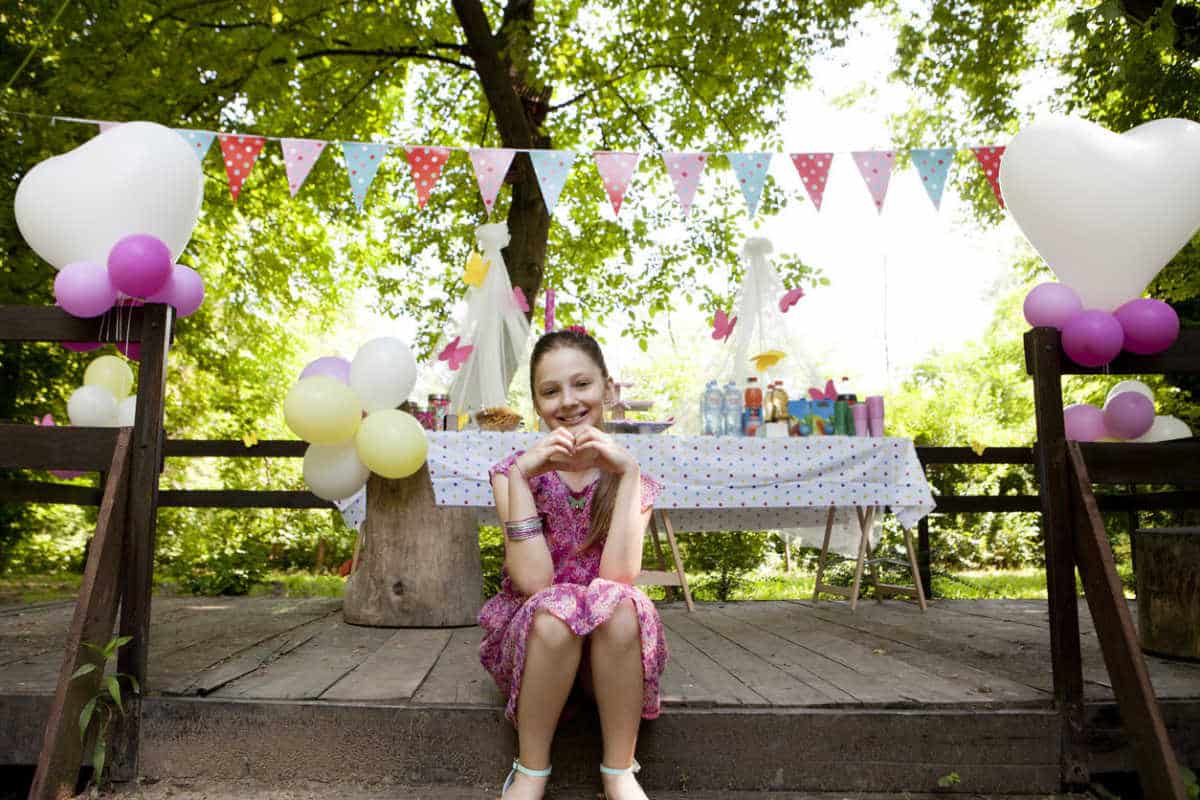 A girl sitting on a wooden bench in front of a table with balloons, celebrating a home away party.