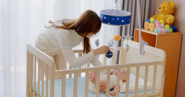 asian mother is playing with cute newborn baby who in crib at home and he watching toys which hanging above cot