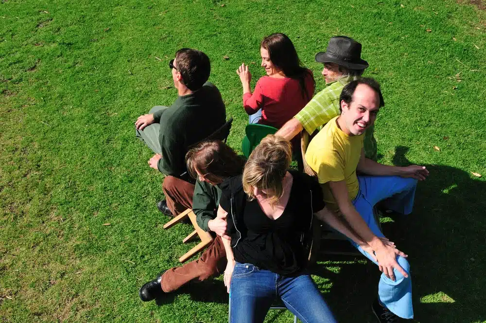 A group of people sitting on a lawn in the sun playing Musical Chairs.